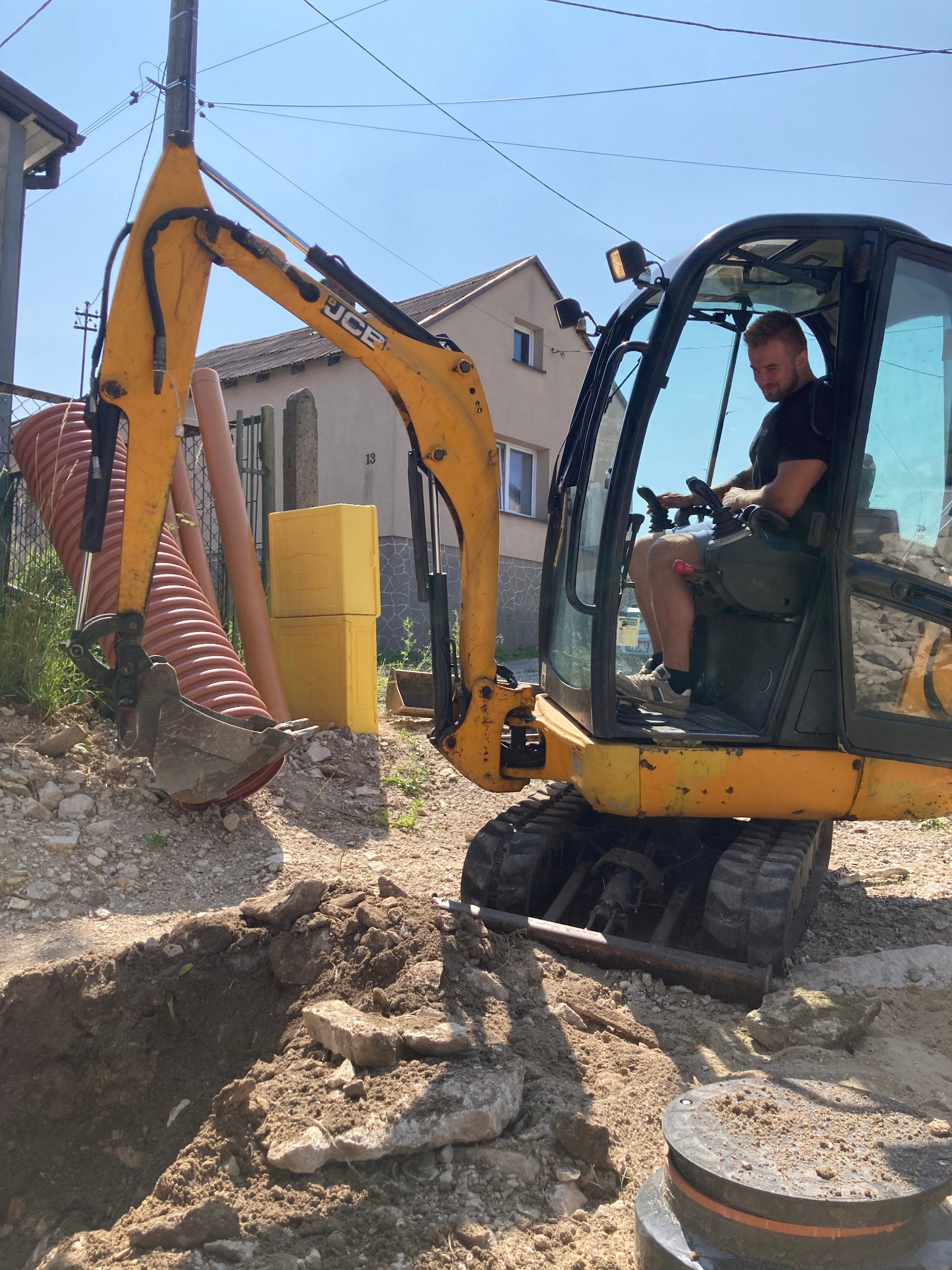 Żółta koparka JCB podczas wykopu rowu pod rury kanalizacyjne, widoczne rury z tworzywa sztucznego i styropianowe płyty izolacyjne, operator w kabinie.
