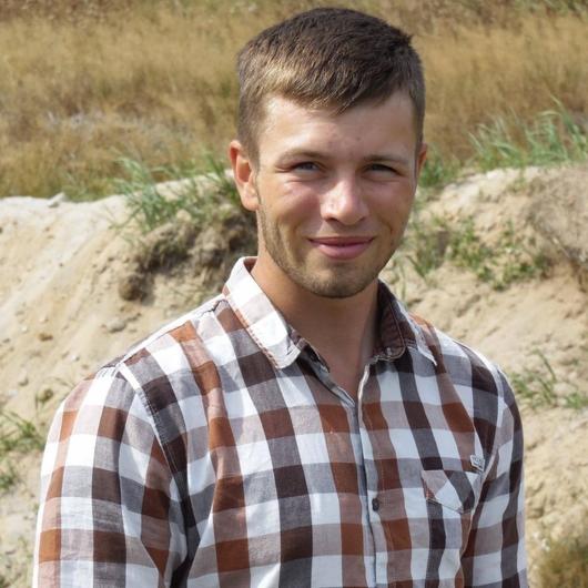 Uśmiechnięty mężczyzna w koszuli w kratę na tle piaszczystego wzgórza i suchej trawy, w naturalnym oświetleniu.