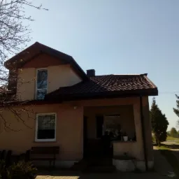 Dwupiętrowy dom z brązowym dachem z dachówki ceramicznej, widok z przodu w słoneczny dzień, drzewo bez liści po lewej stronie.