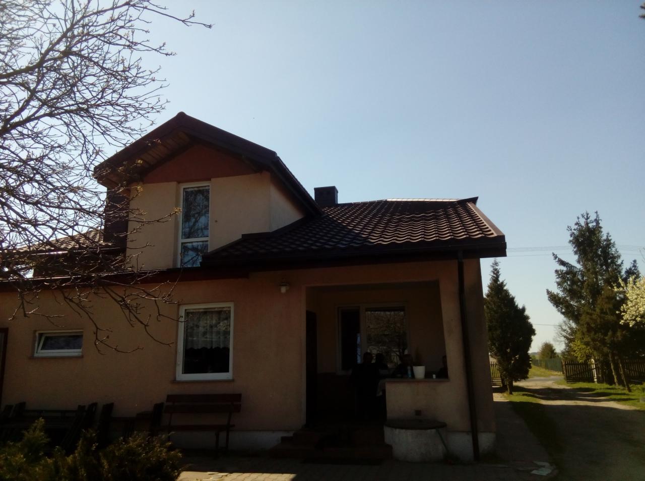 Dwupiętrowy dom z brązowym dachem z dachówki ceramicznej, widok z przodu w słoneczny dzień, drzewo bez liści po lewej stronie.