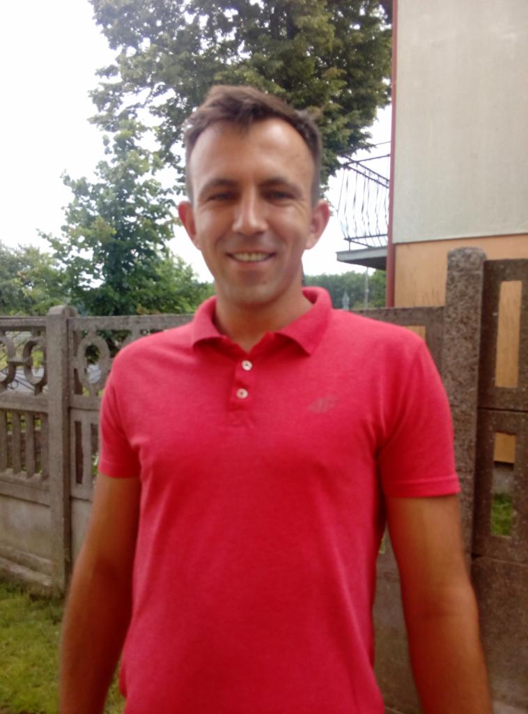 Uśmiechnięty mężczyzna w czerwonej koszulce polo, stojący na tle betonowego ogrodzenia i fragmentu budynku z balkonem.