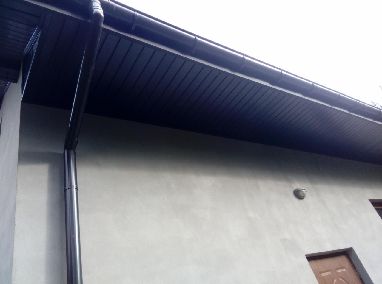 Ciemna podbitka dachowa z rynną spustową zamontowana na jasnej elewacji budynku jednorodzinnego.