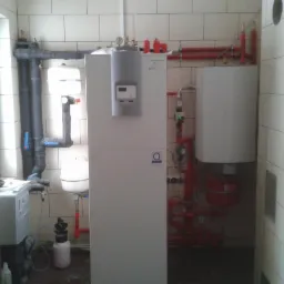 Biała pompa ciepła w kotłowni z widocznymi rurami instalacji grzewczej, zbiornikami wyrównawczymi i elementami sterującymi, na podłodze butelka z płynem.