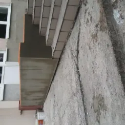 Odnowiona elewacja budynku z białymi oknami, widoczne betonowe schody i świeżo położony tynk. Fragment budynku z oknami i schodami w surowym stanie.