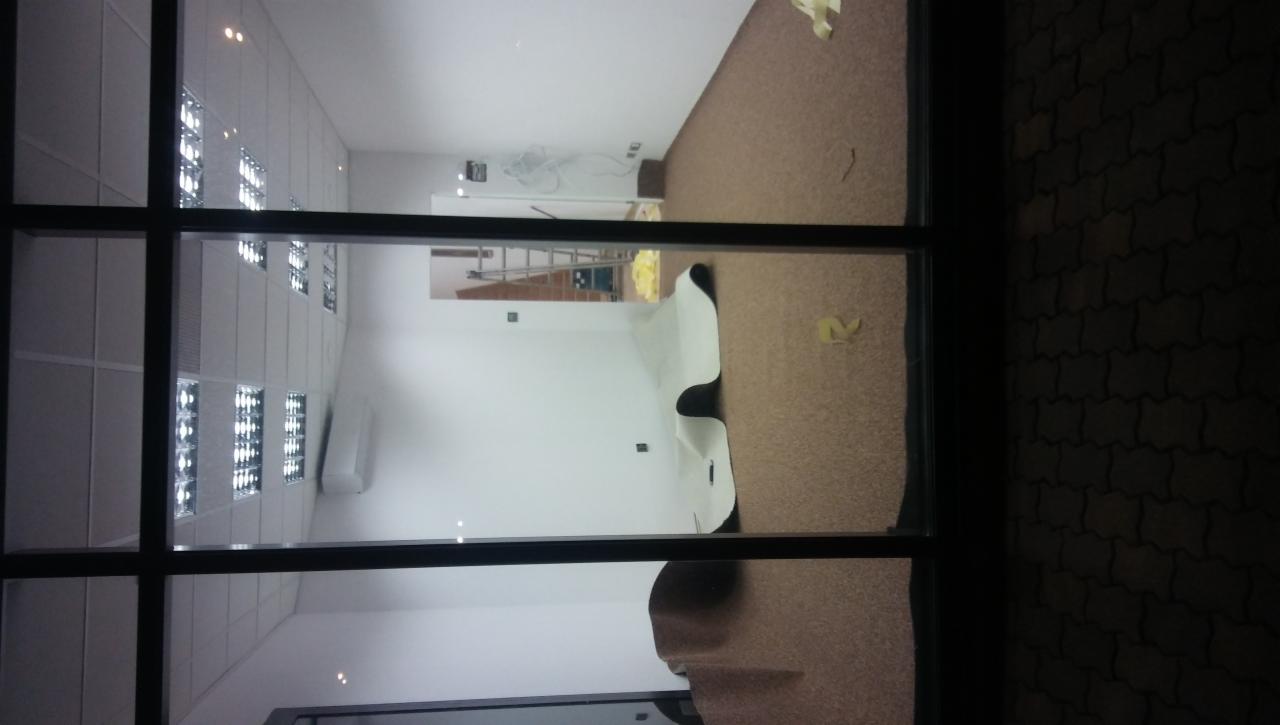 Widok przez okno na wnętrze pomieszczenia w trakcie układania wykładziny podłogowej, z widoczną drabiną i folią ochronną na ścianie.