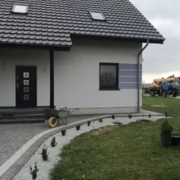 Dom jednorodzinny z nową kostką brukową na podjeździe, obrzeżem z białego grysu, świeżo posadzone tuje, oraz elewacją w odcieniach bieli i szarości z widocznym żurawiem budowlanym w tle.