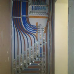 Rozdzielacz instalacji wodnej z licznymi rurami w kolorach niebieskim i miedzianym, zamontowany na ścianie w surowym stanie.