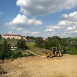 Żółta koparko-ładowarka na piaszczystym, wyrównanym terenie budowy, widoczne ślady opon, w tle budynki mieszkalne i błękitne niebo z obłokami.