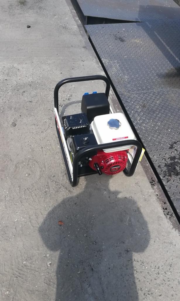 Kompaktowa zagęszczarka gruntu marki Honda, z czerwonym silnikiem i białym zbiornikiem paliwa, stoi na szarym betonie obok metalowej rampy załadunkowej, rzucając cień osoby.