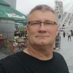 Mężczyzna w okularach robiący selfie na tle kawiarni z parasolem Grolsch i diabelskiego młyna w oddali.