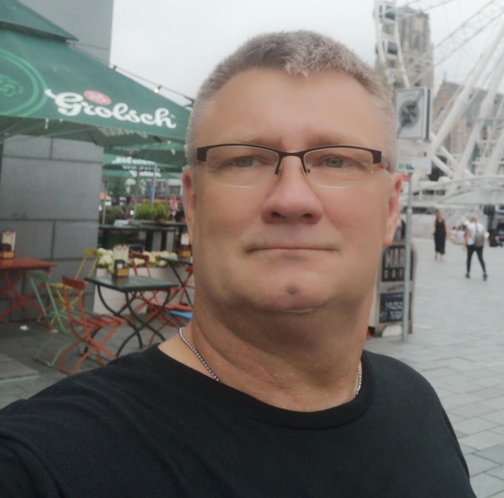 Mężczyzna w okularach robiący selfie na tle kawiarni z parasolem Grolsch i diabelskiego młyna w oddali.