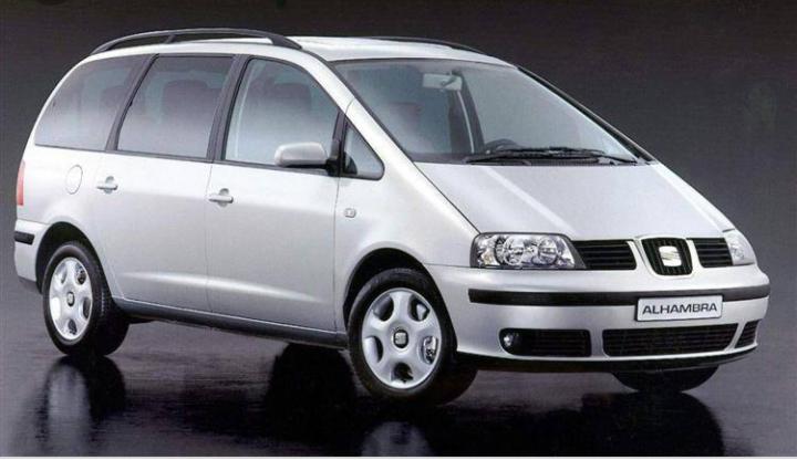 Srebrny minivan Seat Alhambra zaparkowany na błyszczącej, ciemnej powierzchni, prezentowany w studio na jednolitym tle.