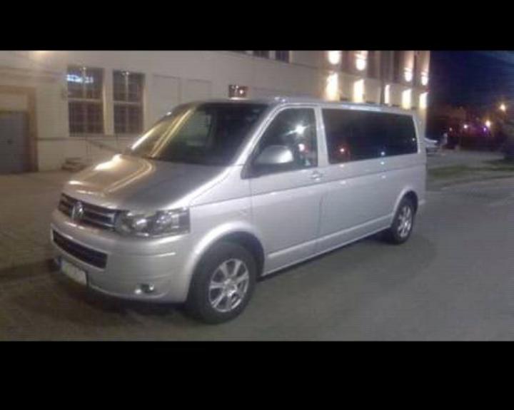 Srebrny Volkswagen Transporter T5 na parkingu miejskim nocą, widoczny z przodu pod kątem, z oświetlonym budynkiem w tle.