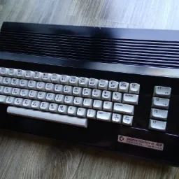 Czarny komputer Commodore 64 z białymi klawiszami na drewnianym blacie. Widoczne detale obudowy i klawiatury, w tym logo Commodore 64 Personal Computer.