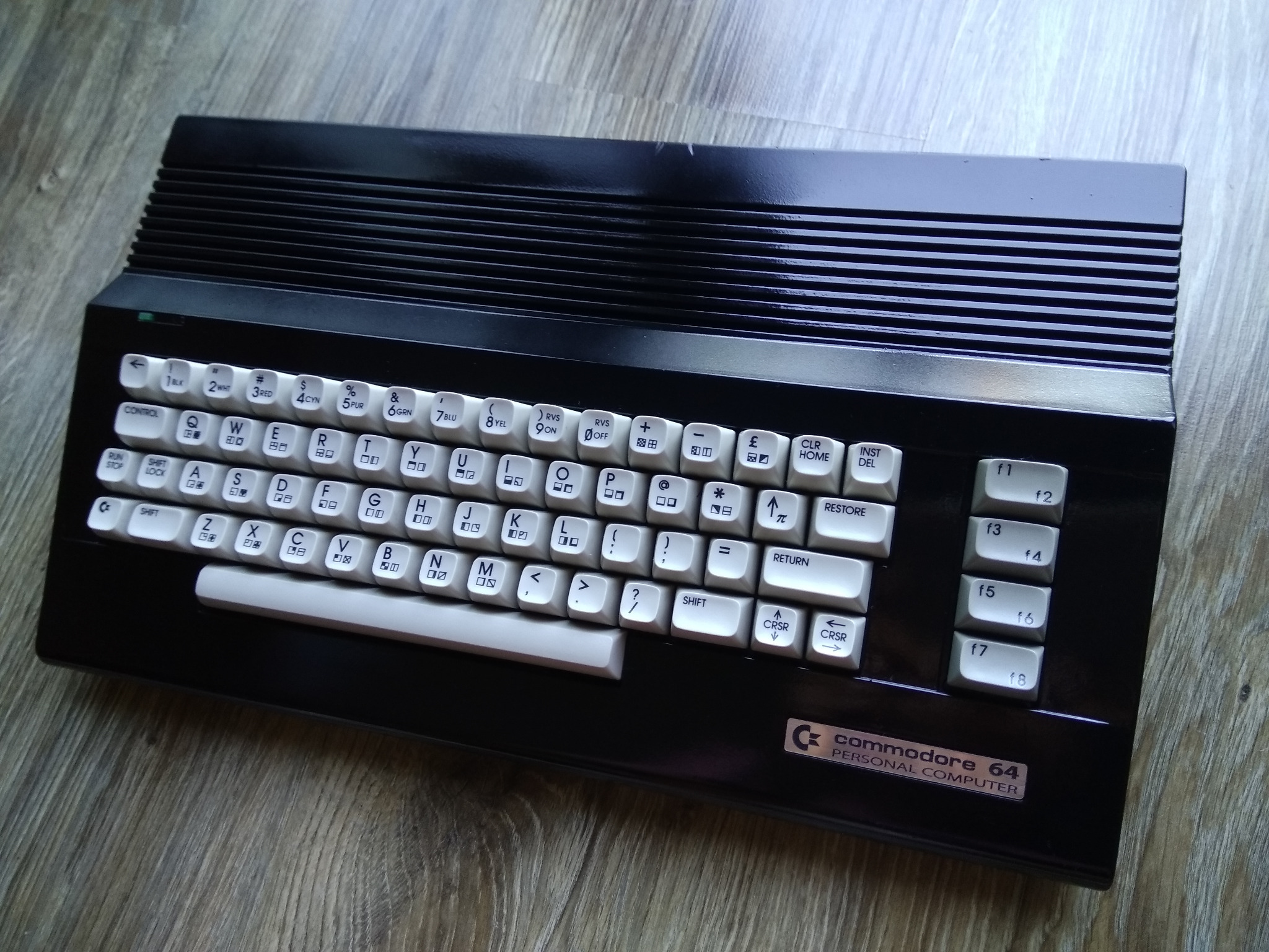 Czarny komputer Commodore 64 z białymi klawiszami na drewnianym blacie. Widoczne detale obudowy i klawiatury, w tym logo Commodore 64 Personal Computer.