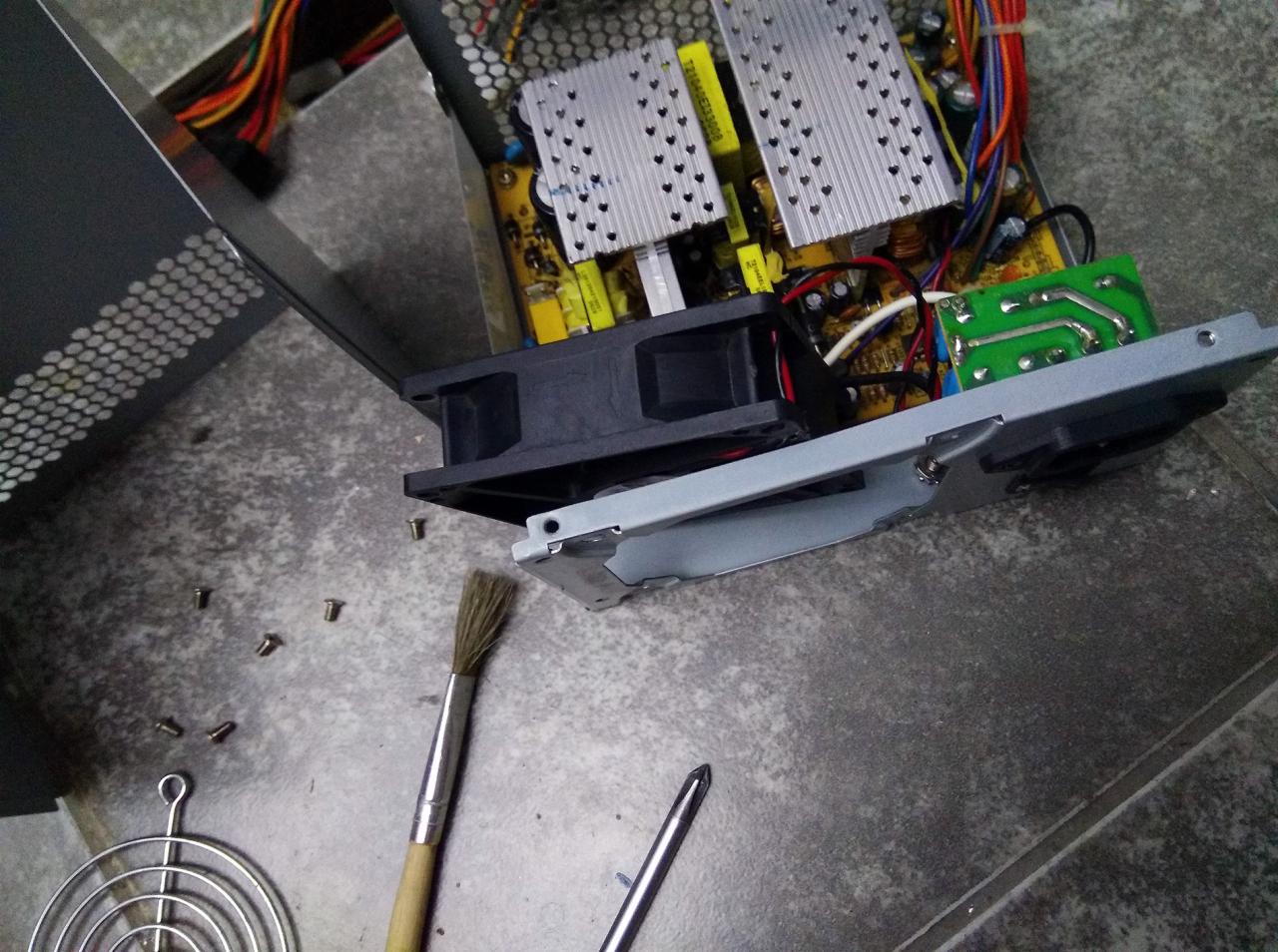 Otwarty zasilacz komputerowy podczas czyszczenia, widoczne radiatory, wentylator, płyta drukowana, śrubki, pędzel do kurzu i śrubokręt na szarym tle.