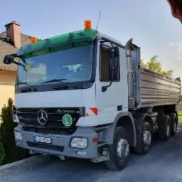 Biała ciężarówka wywrotka Mercedes Actros z pomarańczowymi światłami ostrzegawczymi na dachu, zaparkowana na betonowym podjeździe przed domem jednorodzinnym z czerwonym dachem.