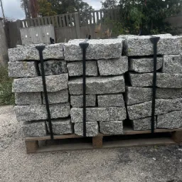 Paleta z ułożonymi warstwami szarej kostki granitowej, zabezpieczona czarnymi pasami, na żwirowym podłożu z widocznym ogrodzeniem i zielenią w tle.