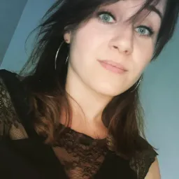 Portret kobiety z zielonymi oczami, srebrnymi kolczykami i czarną koronkową bluzką na tle błękitnej ściany. Ujęcie z dołu, włosy delikatnie rozwiane.