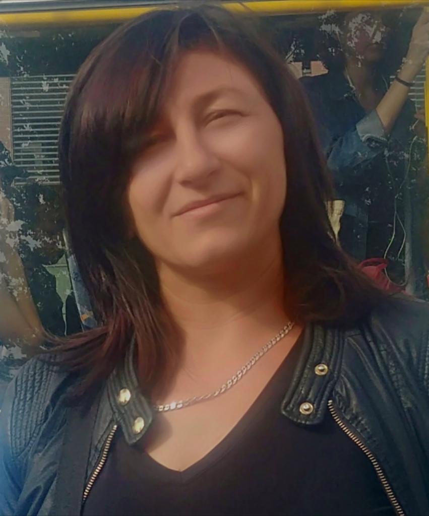 Portret kobiety w skórzanej kurtce, z łańcuszkiem na szyi, na tle wzorzystej ściany. Uśmiech na twarzy, ciemne włosy z grzywką.