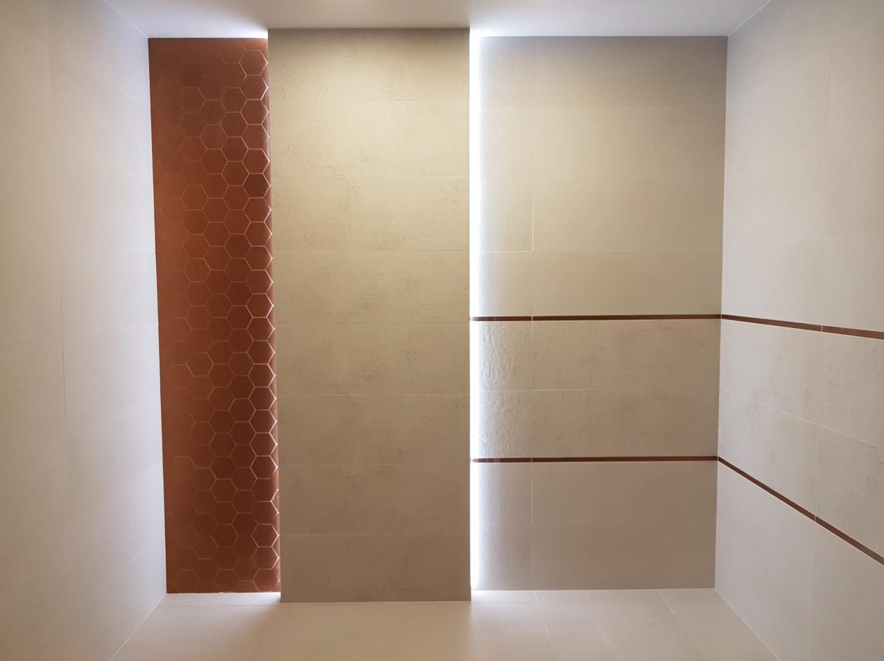 Wykończona łazienka z beżowymi płytkami i pionowym panelem z miedzianymi heksagonami oraz podświetleniem LED w szczelinie między płytkami.