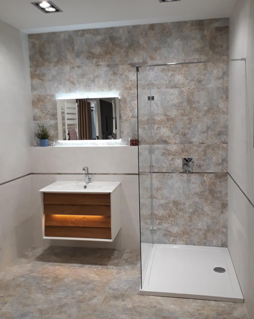 Nowoczesna łazienka z beżowo-szarą mozaiką na ścianach, podświetlanym lustrem, wiszącą szafką pod umywalką z drewnianymi frontami i kabiną prysznicową typu walk-in z chromowanymi okuciami.