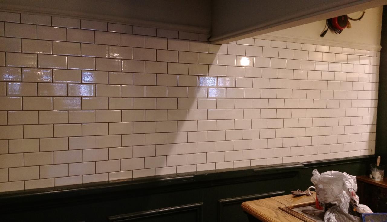 Ściana wyłożona błyszczącymi, beżowymi, prostokątnymi płytkami w stylu retro, z ciemnozieloną boazerią u dołu i widocznymi narzędziami oraz resztkami materiałów po remoncie.