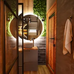 Nowoczesna łazienka z okrągłym lustrem podświetlanym LED, kamienną umywalką nablatową, drewnianymi elementami i ścianą z zielenią wertykalną.