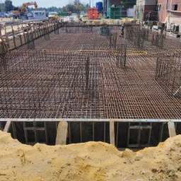 Siatka z prętów zbrojeniowych ułożona w wykopie z drewnianym szalunkiem, przygotowana do zalania betonem, w tle budynek przemysłowy i sprzęt budowlany.