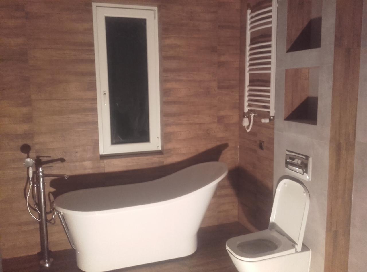 Nowoczesna łazienka z wolnostojącą białą wanną, grzejnikiem drabinkowym i toaletą, wykończona płytkami imitującymi drewno i szarym kamieniem.