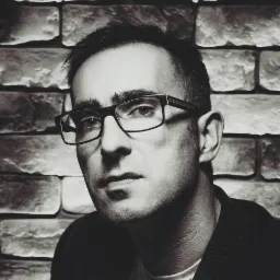 Czarno-biały portret mężczyzny w okularach na tle ceglanej ściany, z oświetleniem podkreślającym rysy twarzy i fakturę cegieł. Wyraz twarzy poważny, skupiony.