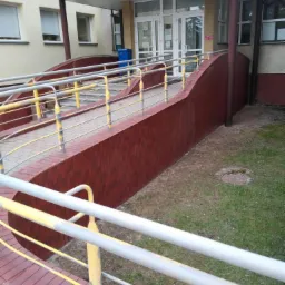 Zewnętrzny podjazd dla niepełnosprawnych z cegły klinkierowej, z metalowymi barierkami pomalowanymi na szaro i żółto, prowadzący do wejścia budynku z białymi drzwiami.