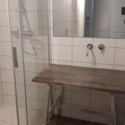 Wykończona łazienka z kabiną prysznicową z przezroczystego szkła i blatem z ciemnego drewna pod umywalką, widoczne białe płytki na ścianach i podłodze.