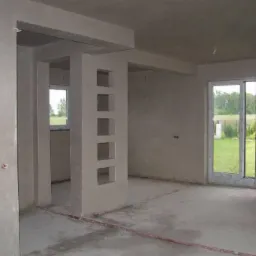 Surowe wnętrze domu w trakcie wykańczania, ściany pokryte świeżym tynkiem, widoczne okno z widokiem na zieleń i filar z dekoracyjnymi wnękami.