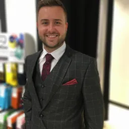 Uśmiechnięty mężczyzna w garniturze w kratę, z bordową poszetką i krawatem w paisley, stoi na tle regału z kolorowymi tkaninami.