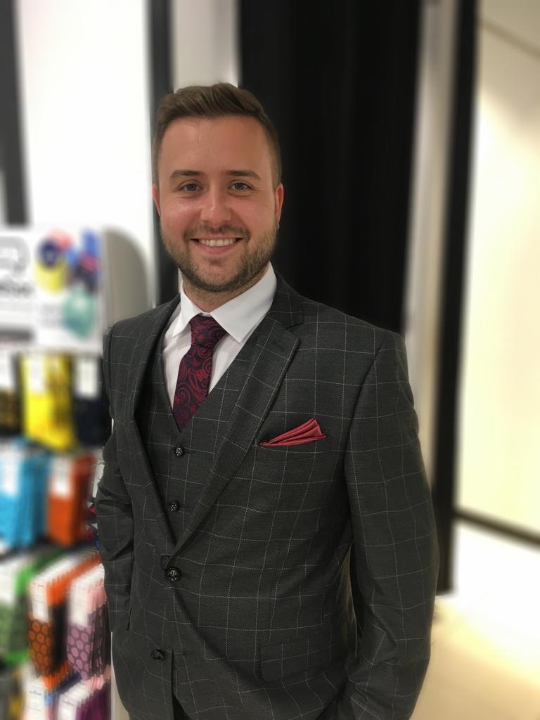 Uśmiechnięty mężczyzna w garniturze w kratę, z bordową poszetką i krawatem w paisley, stoi na tle regału z kolorowymi tkaninami.