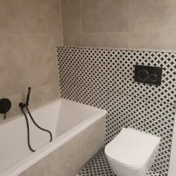Biała wanna z czarną armaturą i biała toaleta w łazience wykończonej szarymi płytkami i dekoracyjną mozaiką w czarno-białe romby.