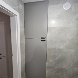 Bud Invest Spółka z Ograniczoną Odpowiedzialnością - Szara szafka w łazience z szarymi płytkami imitującymi marmur, widoczna zza białej futryny drzwi. Minimalistyczny design, nowoczesne wykończenie.