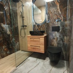 Bud Invest Spółka z Ograniczoną Odpowiedzialnością - Nowoczesna łazienka z kabiną prysznicową typu walk-in, umywalką nablatową na drewnianej szafce i toaletą w kolorze czarnym z marmurkowym wzorem.