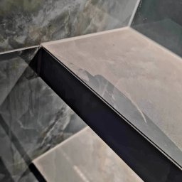 Bud Invest Spółka z Ograniczoną Odpowiedzialnością - Półka z płytek imitujących marmur, wykończona czarną listwą, zamontowana w rogu. Widoczne detale fugi i wzoru płytek. Ujęcie z bliska, podkreślające precyzję wykonania.