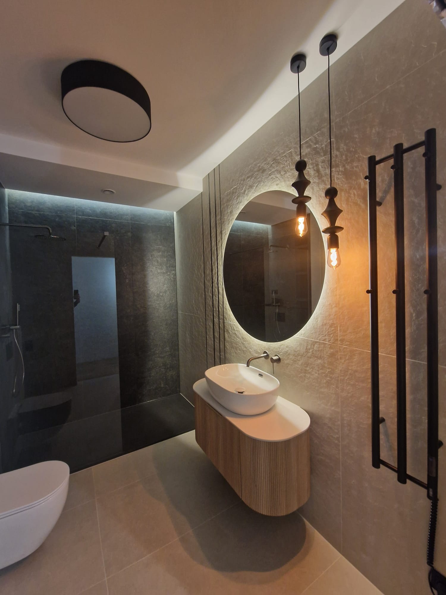 Nowoczesna łazienka z okrągłym lustrem podświetlanym LED, wiszącą szafką z umywalką i grzejnikiem drabinkowym. Minimalistyczny design, szare płytki i oświetlenie dekoracyjne.