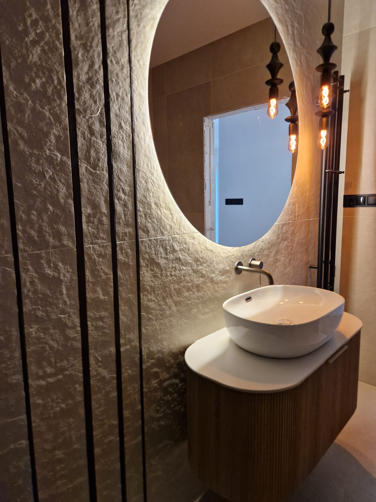 Nowoczesna łazienka: owalne lustro z podświetleniem LED, umywalka nablatowa na szafce z lamelami, dekoracyjna ściana z pionowymi listwami i designerskie lampy wiszące.