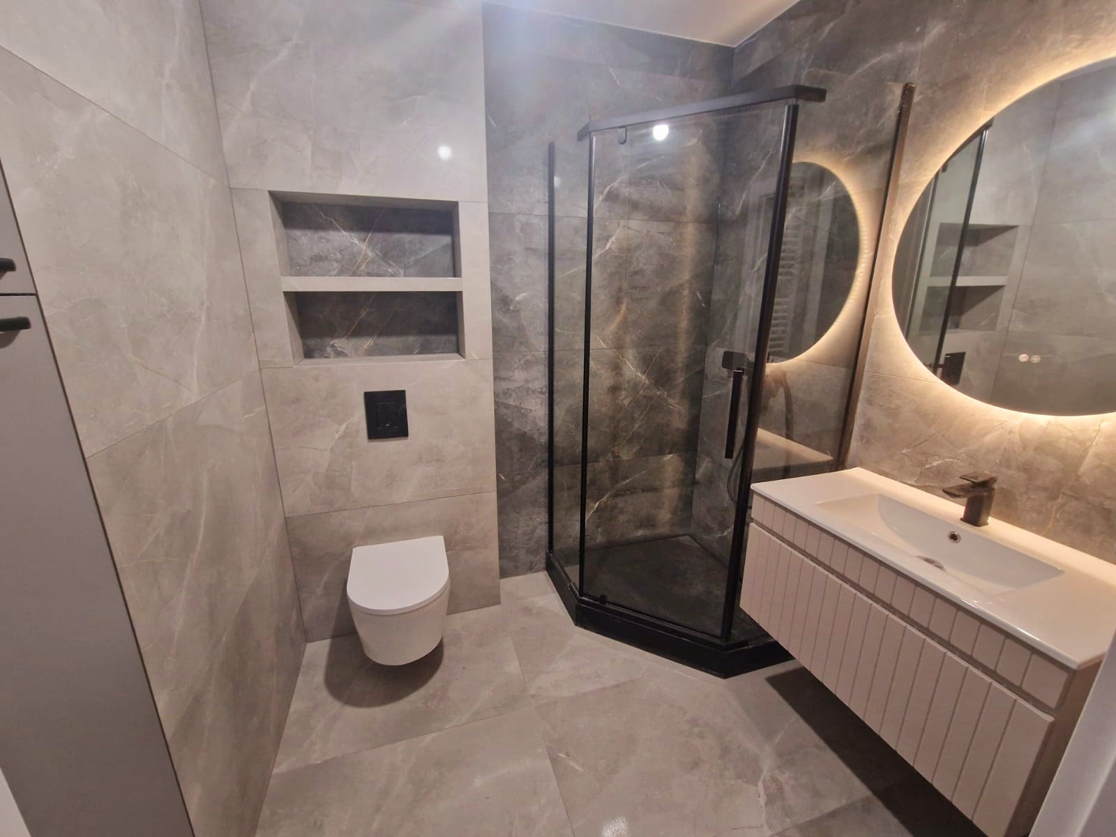 Nowoczesna łazienka z szarymi płytkami imitującymi marmur, kabiną prysznicową z czarnymi profilami i okrągłym lustrem z podświetleniem LED. Minimalistyczny design.