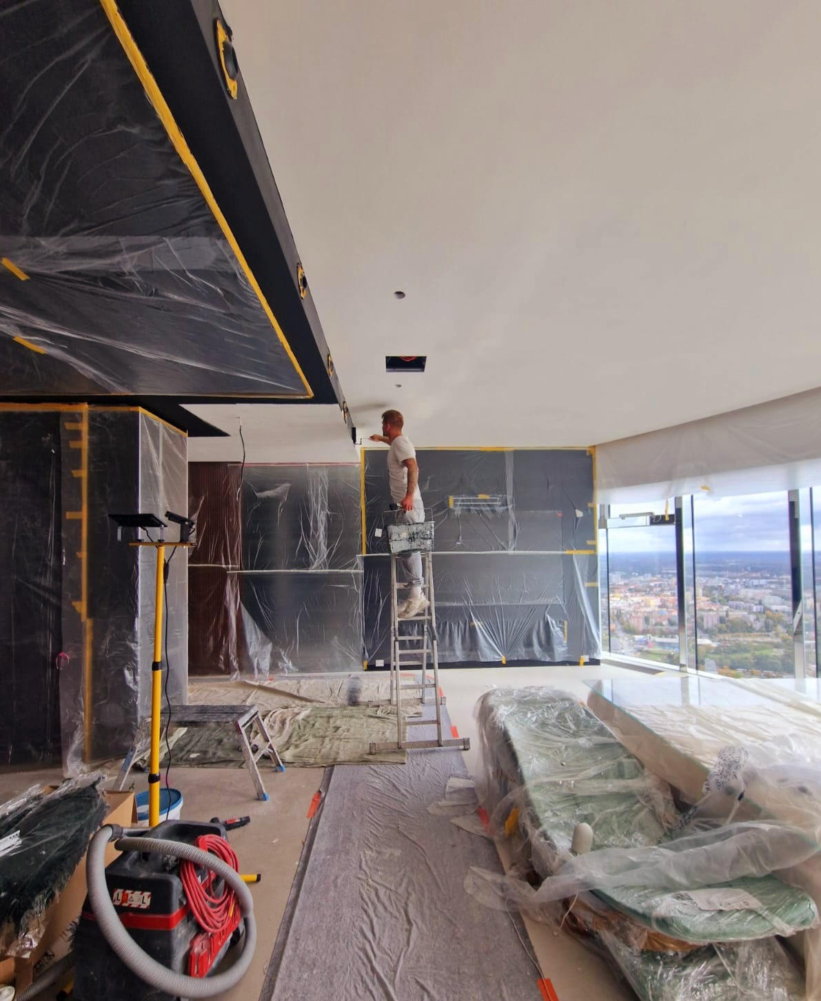 Malarz na drabinie maluje sufit w przestronnym wnętrzu, zabezpieczonym folią. Widoczne narzędzia i panorama miasta za oknem.