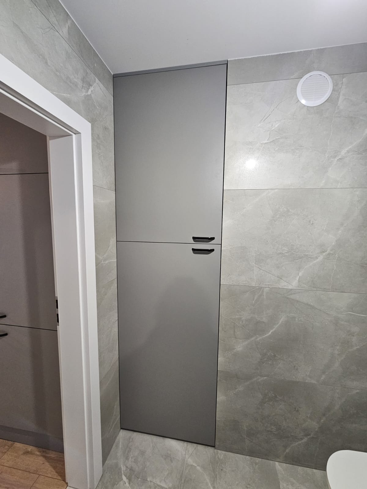 Szara szafka w łazience z szarymi płytkami imitującymi marmur, widoczna zza białej futryny drzwi. Minimalistyczny design, nowoczesne wykończenie.