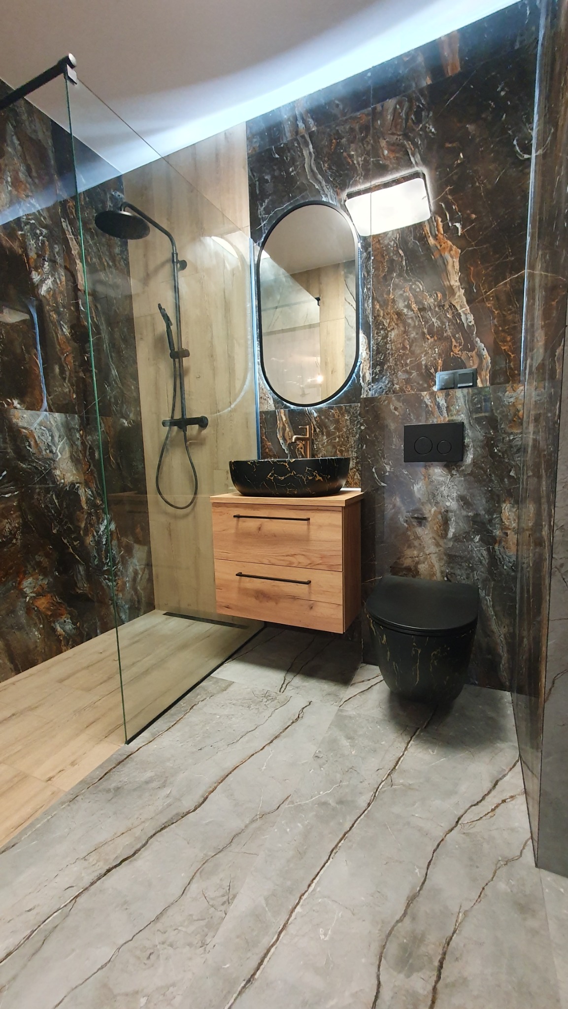 Nowoczesna łazienka z kabiną prysznicową typu walk-in, umywalką nablatową na drewnianej szafce i toaletą w kolorze czarnym z marmurkowym wzorem.