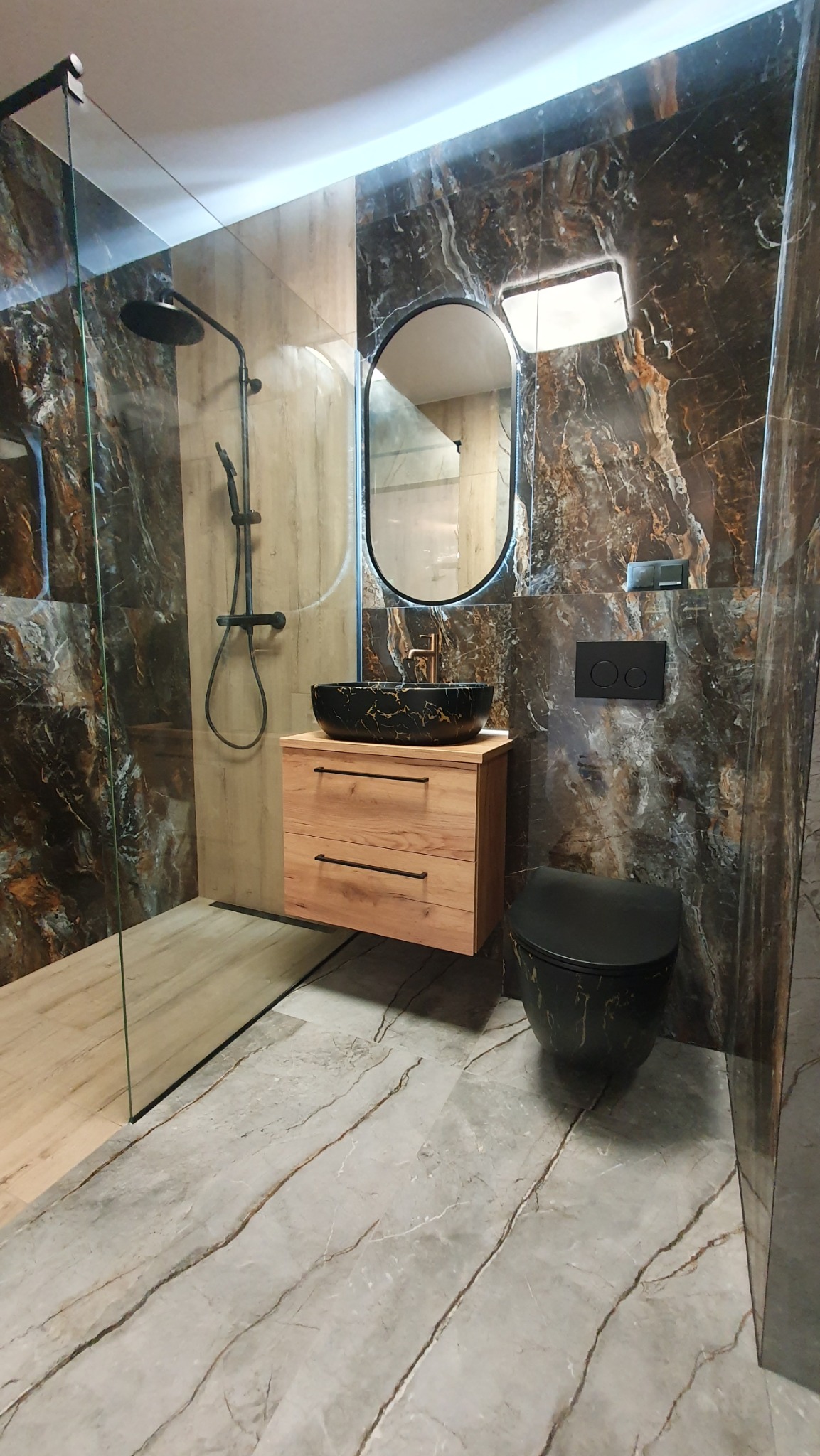 Nowoczesna łazienka z kabiną prysznicową typu walk-in, umywalką nablatową w kolorze czarnym ze złotymi żyłkami i toaletą w tym samym stylu. Marmurowe płytki na ścianach i podłodze.