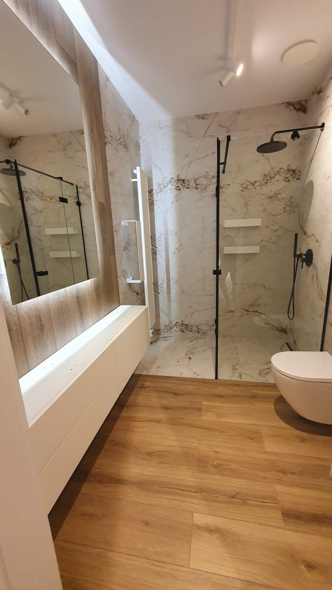 Nowoczesna łazienka z kabiną typu walk-in, marmurowymi płytkami i drewnopodobną podłogą. Uwagę zwraca minimalistyczna armatura w kolorze czarnym i podświetlane lustro.