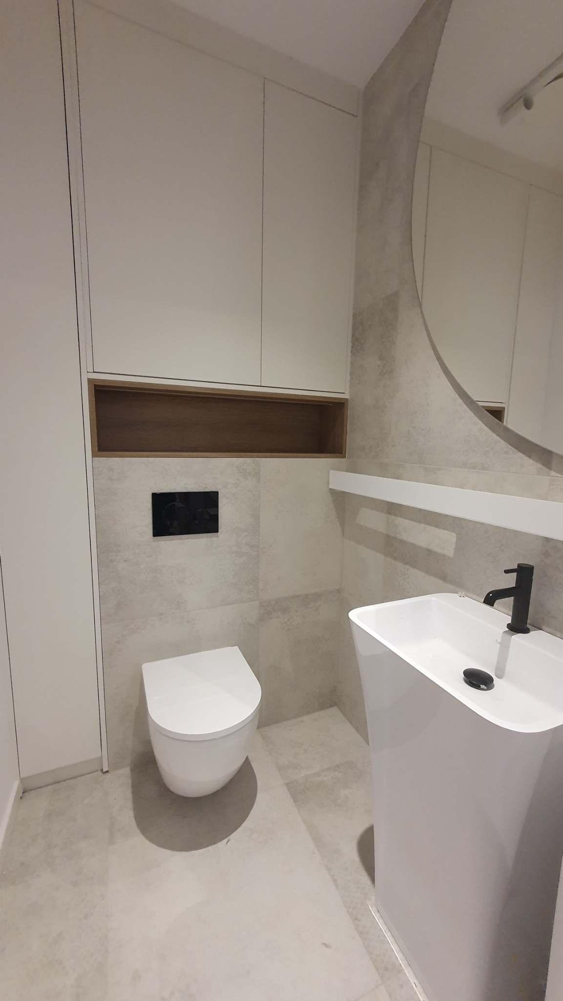 Nowoczesna łazienka w jasnych barwach: podwieszana toaleta, umywalka kolumnowa, duże lustro i szafki w zabudowie. Minimalistyczny design z akcentami drewna.