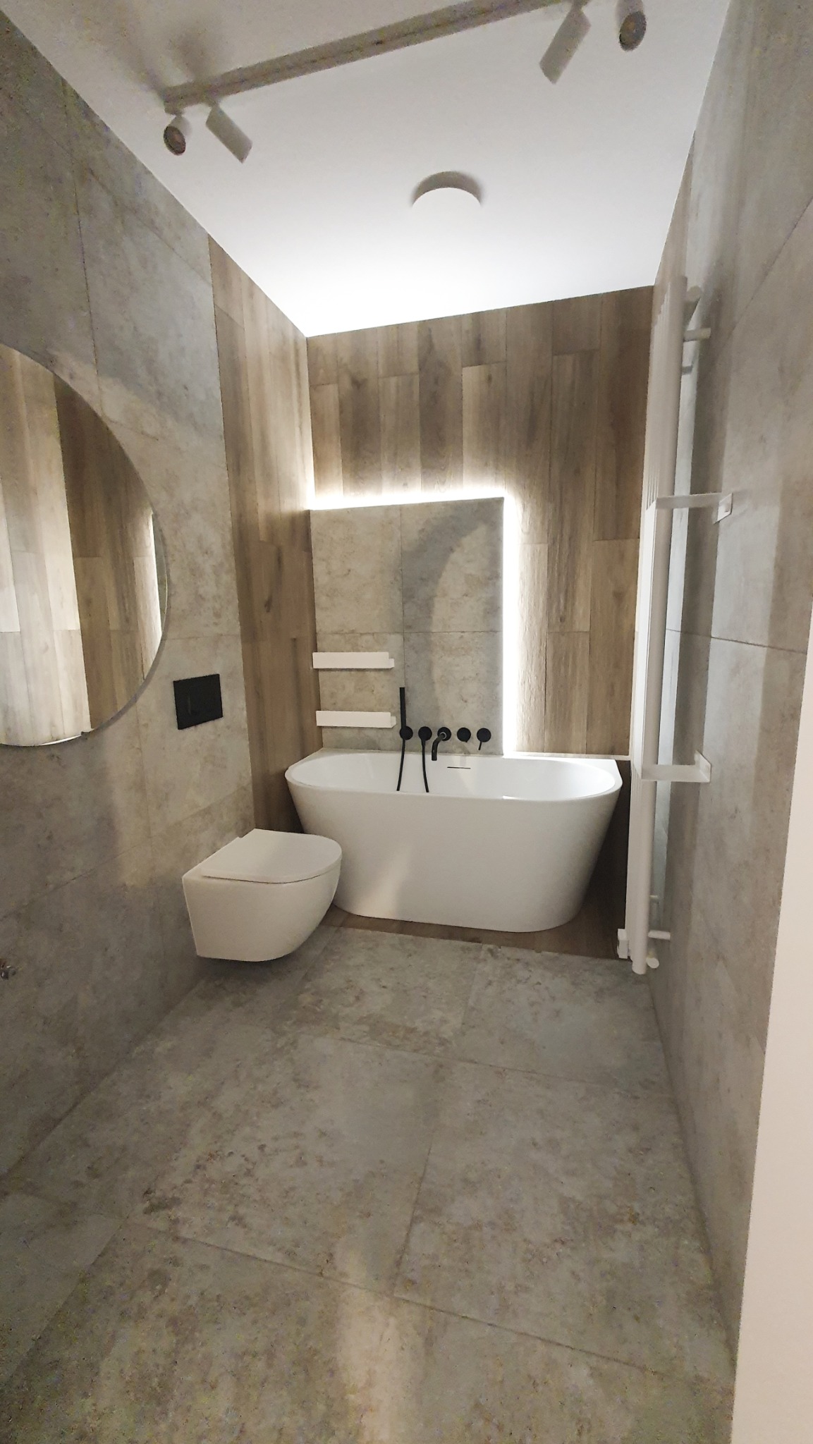 Nowoczesna łazienka z wolnostojącą wanną, podświetleniem LED, wiszącą toaletą i grzejnikiem drabinkowym. Neutralna kolorystyka płytek i drewniane akcenty.